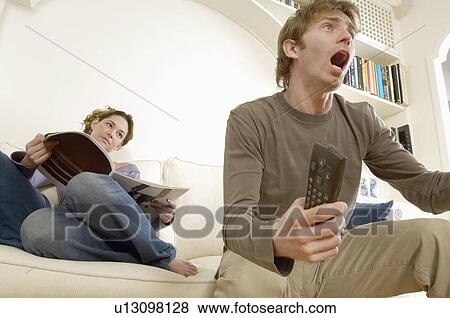 Stock Photo - Young Man Watching Television and Cheering with woman reading magazine behind. Fotosearch