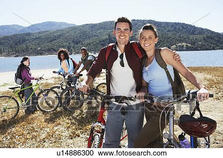 Stock Image - Young people standing with mountain bikes in front of lake. Fotosearch