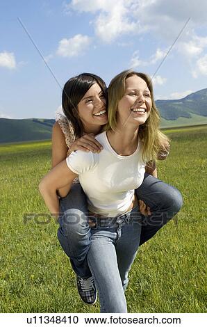 Stock Image - Young woman giving friend piggyback in mountain field, half length. Fotosearch