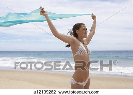 Stock Image - Young woman in bikini holding shawl on beach. Fotosearch