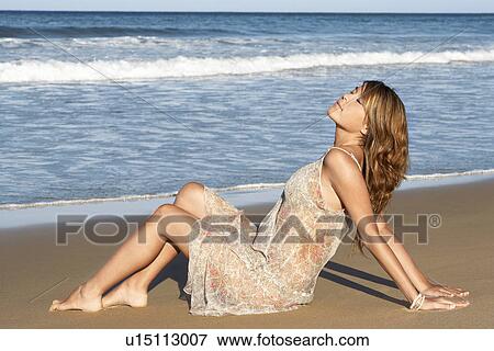 Stock Photo - Young woman in summer dress sitting by sea, side view. Fotosearch