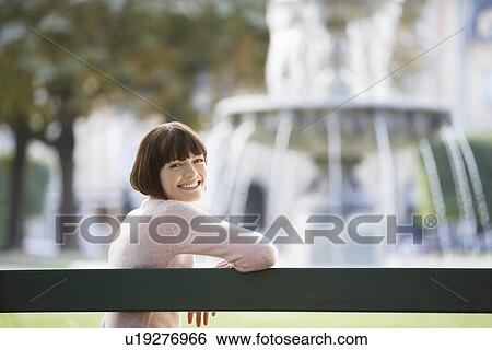 Young woman sitting on bench in front of fountain, looking over shoulder View Large Photo Image Stock Photograph - Young woman sitting on bench in front of fountain, looking over shoulder. Fotosearch