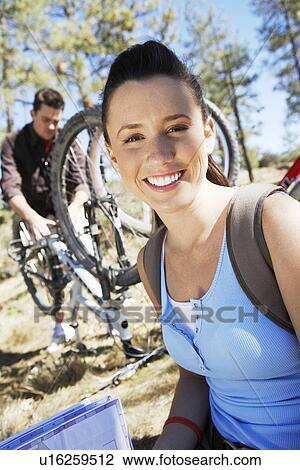Stock Image - Young woman sitting outdoors, man repairing mountain bike in background, portrait.. Fotosearch