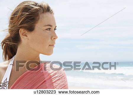 Stock Image - Young woman standing on beach, close-up, head and shoulders. Fotosearch
