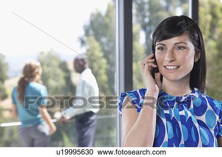 Stock Image - Young woman using mobile phone indoors, smiling. Fotosearch