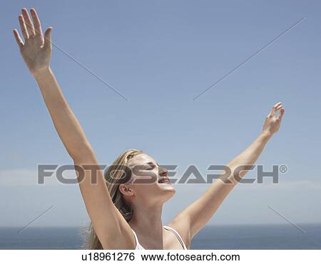 Stock Photograph - Young woman with arms outstretched and eyes closed on beach. Fotosearch
