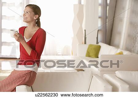 Stock Image - Young woman with drink in living room. Fotosearch