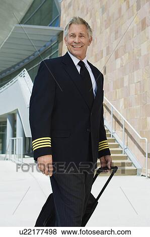Stock Photo - Airline pilot pulling suitcase outside building, portrait. Fotosearch