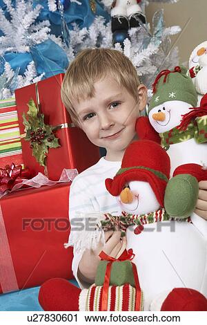 Stock Image - Boy (5-6) holding stuffed snowman in front of Christmas tree, portrait. Fotosearch