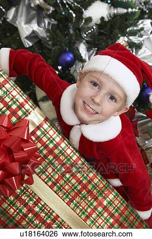 Stock Photograph - Boy (5-6) in Santa costume holding present by Christmas tree. Fotosearch