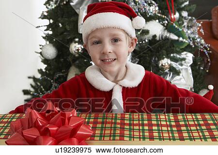 Stock Photography - Boy (5-6) in Santa costume holding present by Christmas tree. Fotosearch