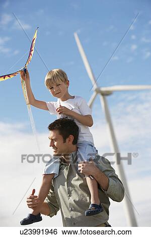 Stock Photograph - Boy (7-9) holding kite, sitting on fathers shoulders at wind farm. Fotosearch