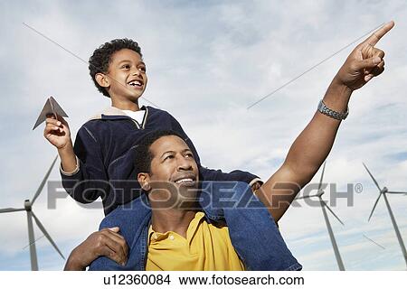 Boy (7-9) holding paper plane, on fathers shoulders at wind farm View Large Photo Image Picture - Boy (7-9) holding paper plane, on fathers shoulders at wind farm. Fotosearch