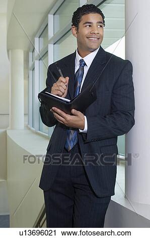 Stock Image - Business man holding diary standing by window in office building. Fotosearch