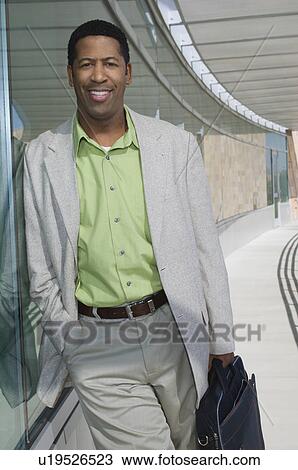 Stock Image - Business man standing by window, outside office building, portrait. Fotosearch