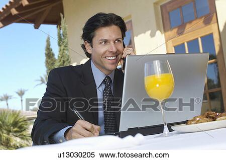Stock Photography - Business man talking on phone and writing in front of laptop, outdoors. Fotosearch