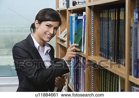 Stock Image - Business woman putting books on shelf in library, portrait. Fotosearch