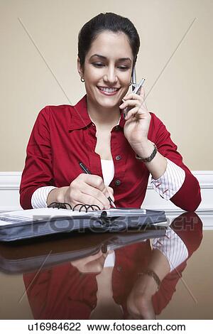Stock Image - Business woman using mobile phone and writing in diary in office. Fotosearch