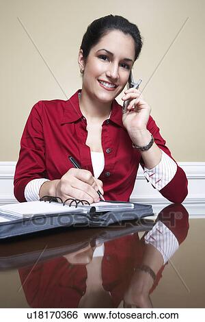 Stock Photograph - Business woman using mobile phone and writing in diary in office, portrait. Fotosearch