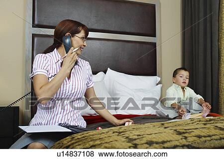 Stock Photo - Business woman using phone with son on bed. Fotosearch