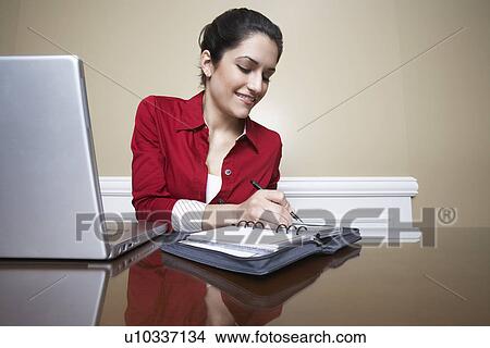 Picture - Business woman writing in diary beside laptop in office. Fotosearch