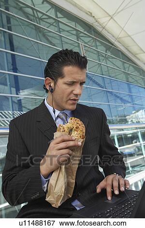 Stock Photo - Businessman eating bagel while using laptop with earpiece in outside office building. Fotosearch