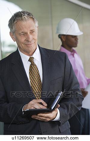 Businessman holding day planner with construction worker behind View Large Photo Image Stock Photo - Businessman holding day planner with construction worker behind. Fotosearch