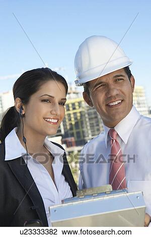 Stock Image - Businessman in hardhat showing clipboard to businesswoman with earpiece. Fotosearch