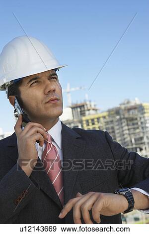Stock Photo - Businessman in hardhat using mobile phone, outdoors. Fotosearch