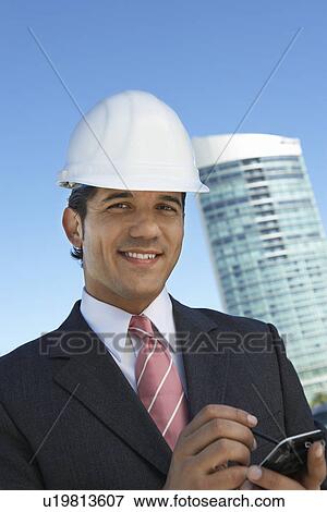 Stock Photo - Businessman in hardhat using PDA outdoors, portrait. Fotosearch