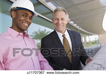 Stock Photo - Businessman looking at blue prints with construction worker, portrait. Fotosearch
