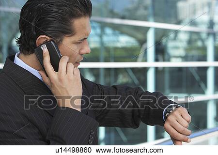Businessman using mobile phone and checking watch outside office building View Large Photo Image Stock Image - Businessman using mobile phone and checking watch outside office building. Fotosearch