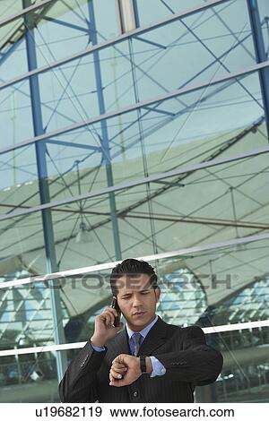 Stock Photo - Businessman using mobile phone and checking watch outside office building. Fotosearch