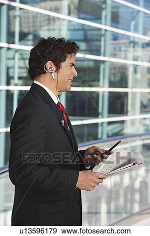 Businessman using mobile phone, reading newspaper outside office building View Large Photo Image Stock Photo - Businessman using mobile phone, reading newspaper outside office building. Fotosearch