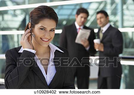Stock Image - Businesswoman using earpiece with businessmen in background. Fotosearch