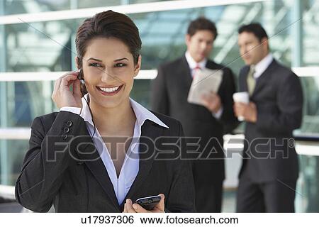 Businesswoman using earpiece with businessmen in background View Large Photo Image Stock Photograph - Businesswoman using earpiece with businessmen in background. Fotosearch