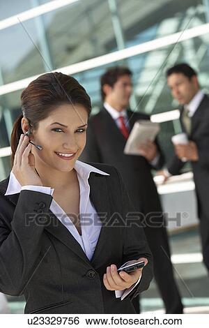 Businesswoman using earpiece with businessmen in background View Large Photo Image Stock Photograph - Businesswoman using earpiece with businessmen in background. Fotosearch