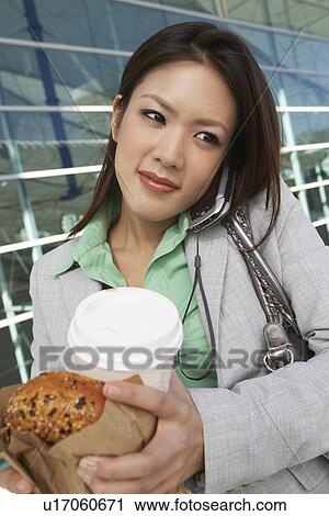 Stock Image - Businesswoman using mobile phone holding food and drink, outside office building. Fotosearch
