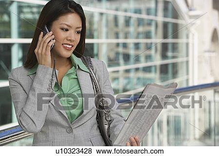 Stock Photo - Businesswoman using mobile phone, reading newspaper outside office building. Fotosearch