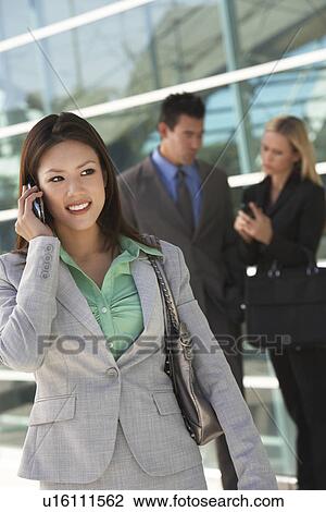 Stock Image - Businesswoman using mobile phone with colleagues in background, outdoors. Fotosearch