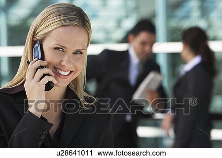Stock Image - Businesswoman using mobile phone with colleagues in background, outdoors. Fotosearch