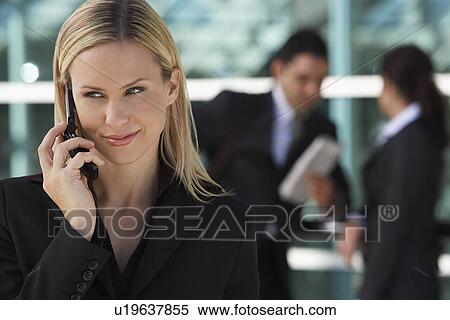 Stock Photography - Businesswoman using mobile phone with colleagues in background, outdoors. Fotosearch