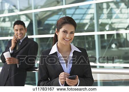 Stock Photo - Businesswoman using PDA with businessman using mobile phone in background, outdoors. Fotosearch