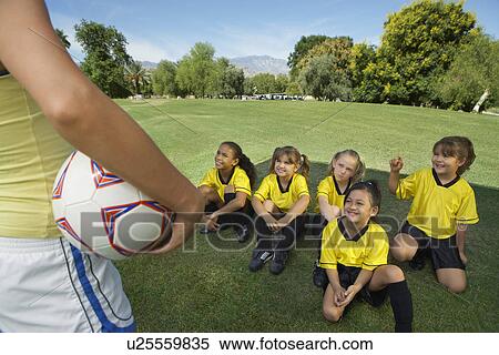 Stock Photography - Coach standing in front of group of girl soccer players (7-9 years) sitting on lawn, elevated view. Fotosearch
