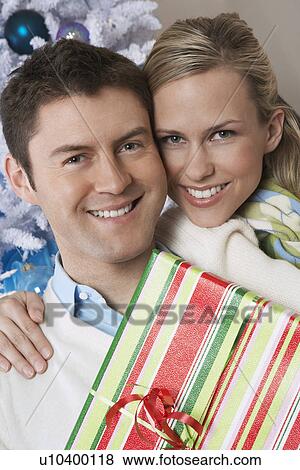 Couple holding present by Christmas tree, portrait View Large Photo Image Stock Photo - Couple holding present by Christmas tree, portrait. Fotosearch