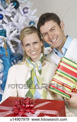 Couple holding presents by Christmas tree, portrait View Large Photo Image Stock Photo - Couple holding presents by Christmas tree, portrait. Fotosearch