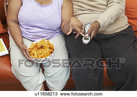 Stock Image - Couple sitting on couch with crisps and phone, mid section. Fotosearch