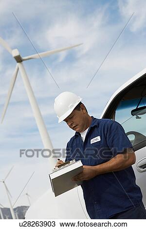 Stock Image - Engineer writing on clipboard by car at wind farm. Fotosearch