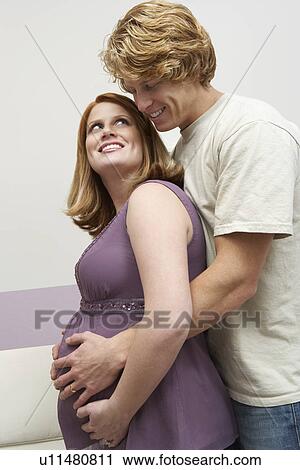 Stock Image - Expectant couple embracing at home, portrait. Fotosearch