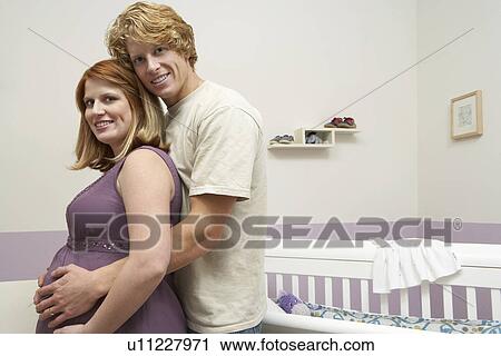 Stock Image - Expectant couple embracing by cradle, portrait. Fotosearch